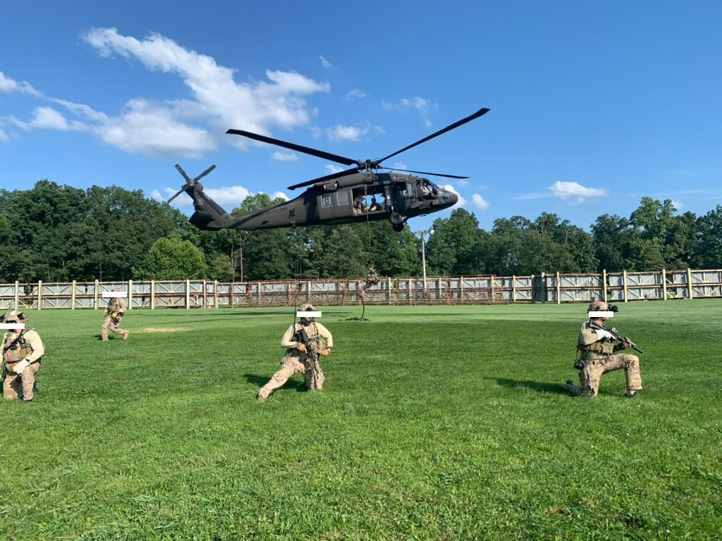 Military training exercise with soldiers on the ground and a helicopter hovering overhead, showcasing tactical equipment and safety gear in an outdoor field environment.